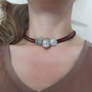 以下が含まれることがあります： 3つの銀色の装飾的な丸いペンダントが付いた、茶色の革製のチョーカーネックレスです。
