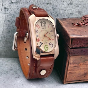 May include: A brown leather wristwatch with a rose gold oval-shaped face. The watch face has a white background with colourful numbers and a small second hand. The watch is on a brown leather strap with a buckle fastening.