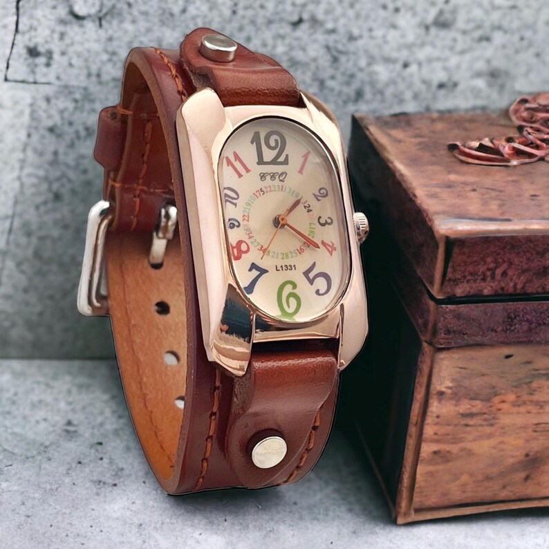 May include: A brown leather wristwatch with a rose gold oval-shaped face. The watch face has a white background with colourful numbers and a small second hand. The watch is on a brown leather strap with a buckle fastening.