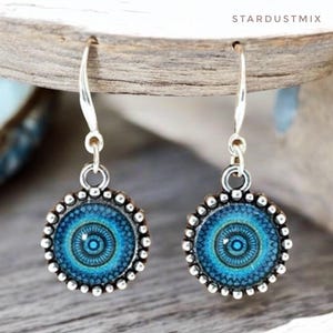 May include: A pair of silver-toned drop earrings featuring circular pendants with a blue and teal mandala-style pattern. The edges of the circular settings are decorated with a beaded texture. The earrings hang from silver-toned metal hooks.