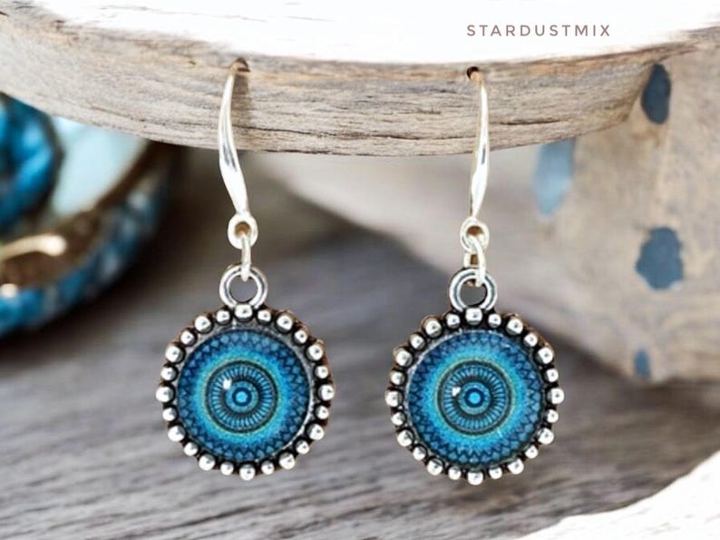 May include: A pair of silver-toned drop earrings featuring circular pendants with a blue and teal mandala-style pattern. The edges of the circular settings are decorated with a beaded texture. The earrings hang from silver-toned metal hooks.