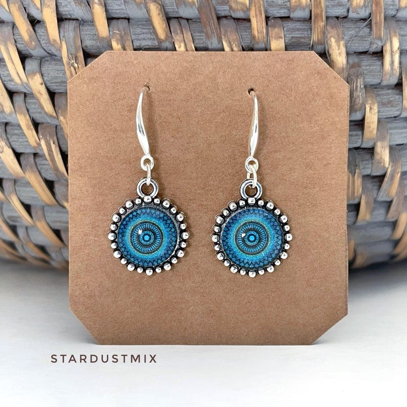 May include: A pair of silver-toned drop earrings featuring circular pendants with a blue and black mandala-style pattern. The edges of the pendants are detailed with a beaded texture, and the earrings hang from simple silver-toned shepherd hooks.