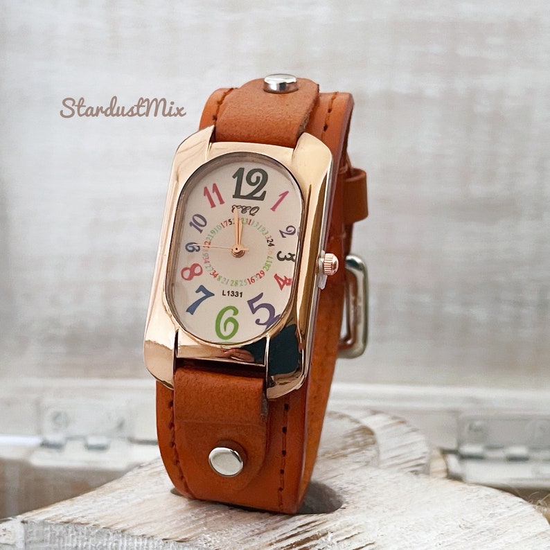 May include: A brown leather wristwatch with a rose gold case and a white face. The numbers on the face are in different colors. The watch has a rectangular face and a unique design.