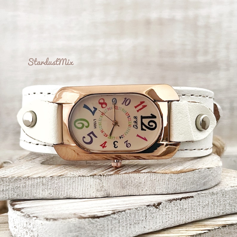 May include: White leather wristwatch with a rose gold case and a colourful face. The watch face has a white background with black hands and colourful numbers.