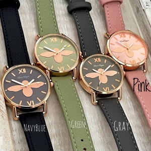 May include: Three wristwatches with different colored leather bands: navy blue, green, and pink. Each watch has a gold-tone case with a bee design on the face and Roman numeral markings. The text "Ban Diya" is printed on the face of each watch.