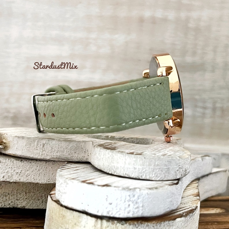 May include: A rose gold watch with a light green leather band. The watch is on a white wooden surface.