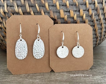 May include: Two pairs of silver earrings on brown card stock. The earrings on the left are oval shaped with a textured surface. The earrings on the right are round with a textured surface. The card stock has the text "Stardustmix" printed on the bottom.