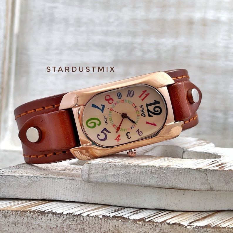 May include: A rose gold watch with a brown leather strap. The watch face is oval shaped and has colourful numbers. The watch face also has the word "Dodo" printed on it.