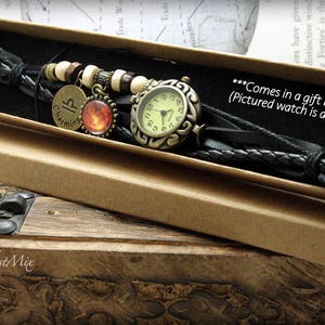 Puede incluir: Un brazalete de reloj de cuero negro con una esfera de reloj dorada y un dije con el s&iacute;mbolo astrol&oacute;gico de Piscis. El brazalete est&aacute; en una caja de regalo marr&oacute;n.