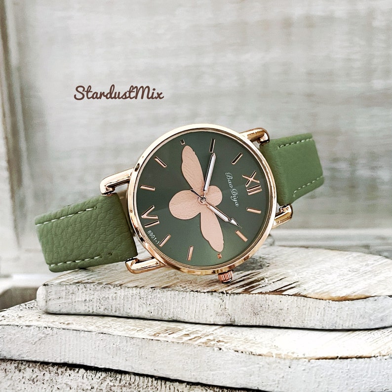 May include: A green leather wristwatch with a rose gold case and a bee design on the face. The watch face has Roman numeral hour markers and the text "Bao-Diga" at the 6 o'clock position.