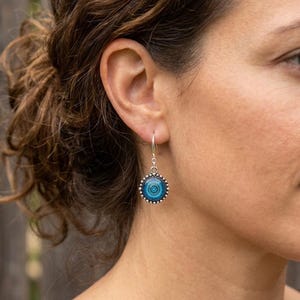 May include: A silver drop earring featuring a circular pendant with a beaded edge. The center of the pendant displays a gradient of blue tones in a concentric circular pattern, suspended from a simple silver hook wire that passes through the ear.
