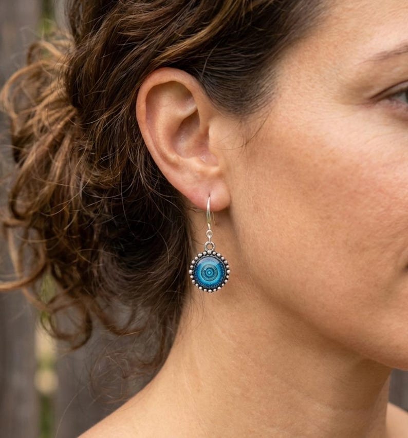 May include: A silver drop earring featuring a circular pendant with a beaded edge. The center of the pendant displays a gradient of blue tones in a concentric circular pattern, suspended from a simple silver hook wire that passes through the ear.
