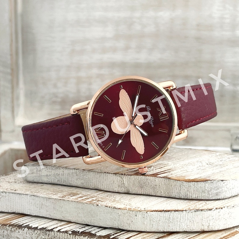 May include: A red leather wristwatch with a rose gold case and a bee design on the face. The watch face has Roman numeral markings and the text "Rose Type" on the face.