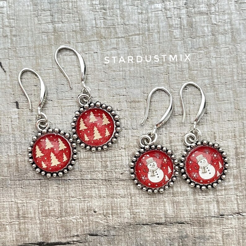 May include: Two pairs of silver-toned drop earrings featuring circular pendants with a red background. One pair displays a pattern of cream-colored pine trees, while the other pair features a white snowman with a scarf. The text STARDUSTMIX is visible.