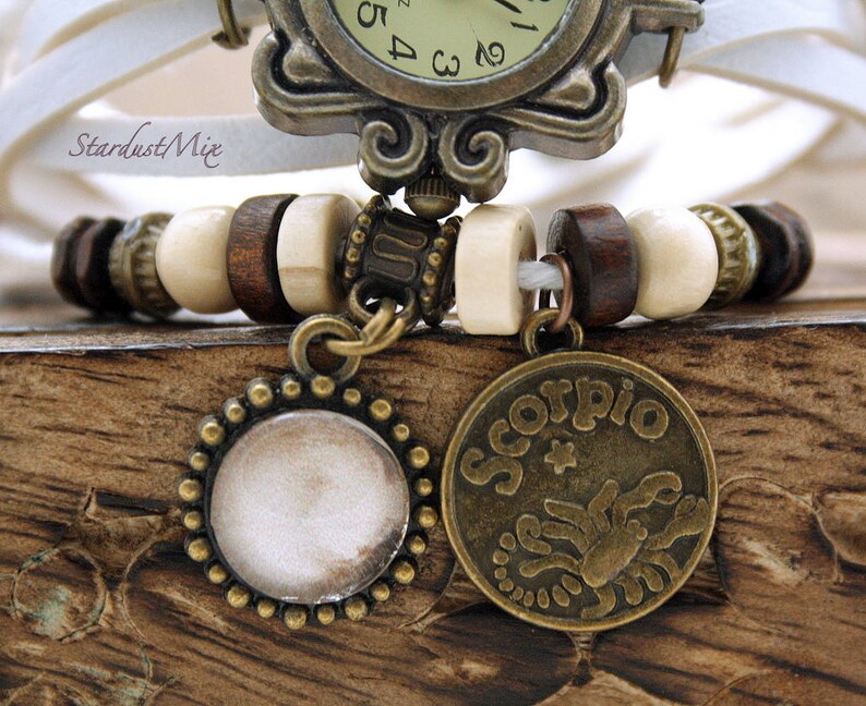 Puede incluir: Un brazalete de reloj de cuero blanco con cuentas de madera marr&oacute;n y dos dijes de bronce. Un dije es un disco redondo, en blanco y el otro es un dije redondo con la palabra "Scorpio" y un dise&ntilde;o de escorpi&oacute;n.