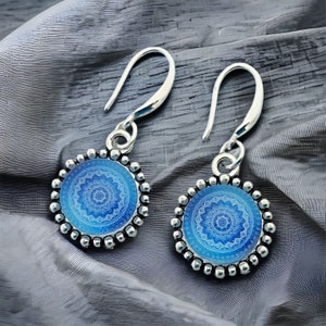 May include: A pair of silver drop earrings featuring circular pendants with a blue and white mandala-style pattern. The edges of the circular settings are decorated with a beaded texture, and the earrings hang from simple silver shepherd hooks.