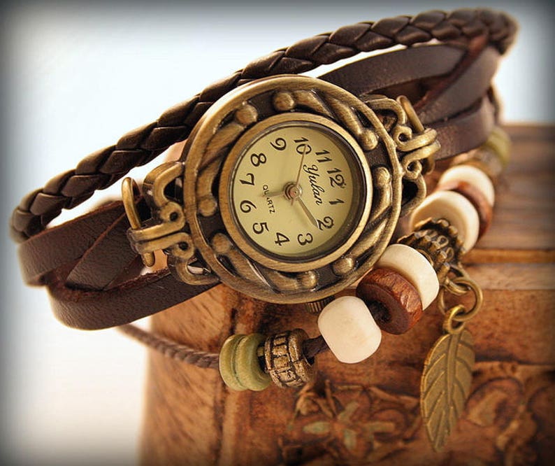 May include: A brown leather wristwatch with a gold-toned face and a beaded charm. The watch face has Roman numerals and the word "Yudan" printed on it. The watch is accented with a brown leather strap and a beaded charm with a feather pendant.
