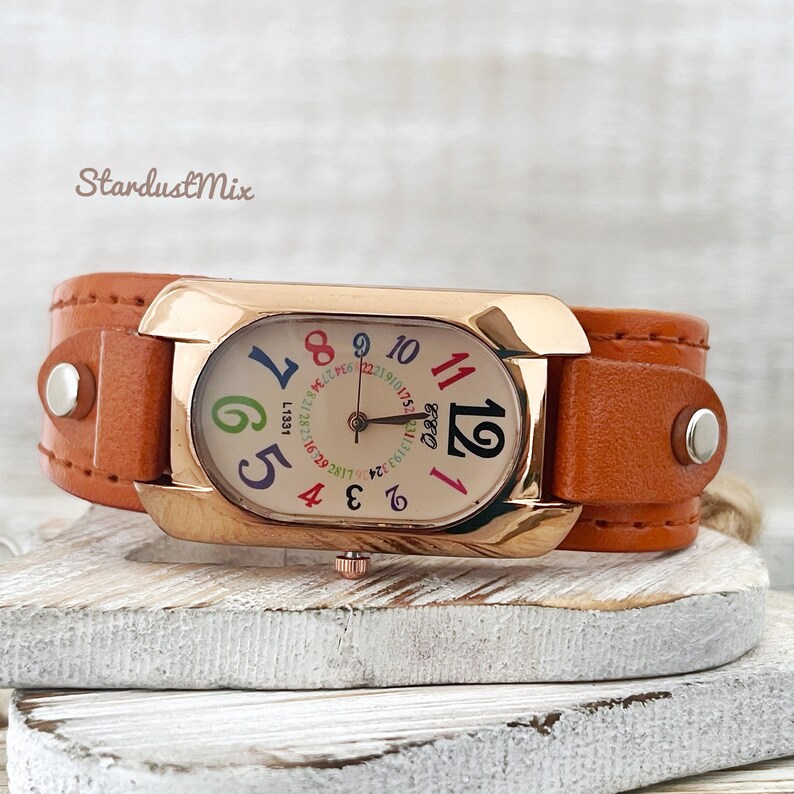 May include: A rose gold watch with an orange leather band. The watch face has a white background with colorful numbers and a black clock hand.