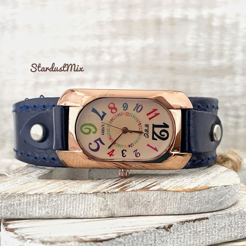 May include: A rose gold watch with a white face and colourful numbers. The watch has a blue leather strap with white stitching.
