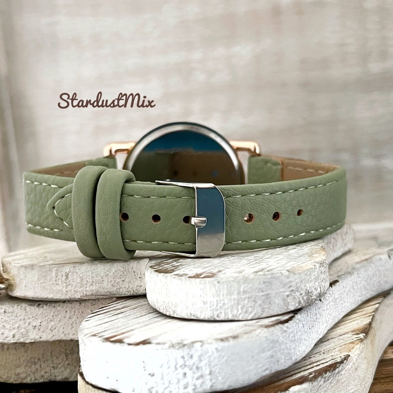 May include: A green leather watch band with a silver buckle. The watch band is on a white wooden surface.