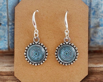 May include: A pair of silver earrings with blue and white mandala designs. The earrings are on a brown card.