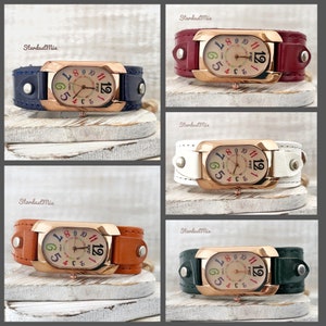 May include: Four different coloured leather wristwatches with rose gold cases and colourful numbers on the face. The watches are on a white wooden background.