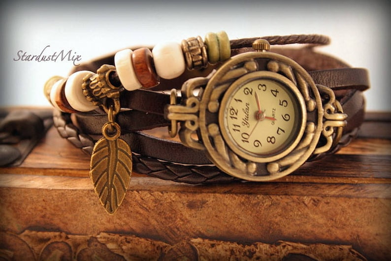 May include: A brown leather wrap watch with a gold-tone face and a feather charm. The watch face has white numbers and a gold-tone Yuluen Quartz inscription.