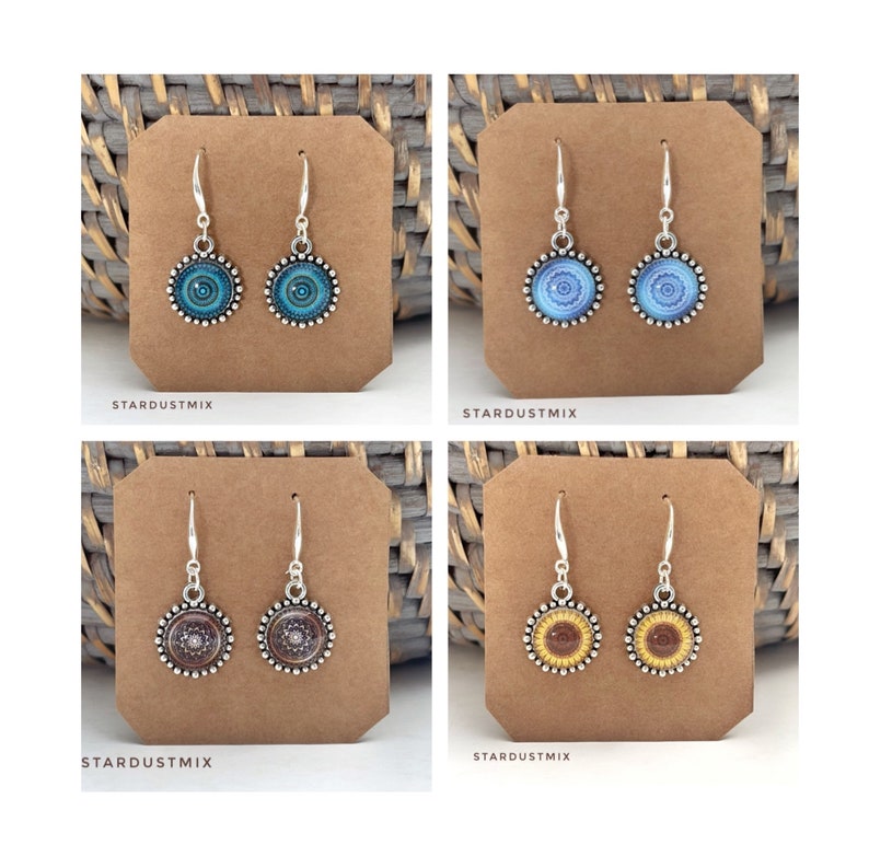 May include: Four pairs of silver-toned drop earrings displayed on brown cardboard cards. Each pair features a circular pendant with a detailed mandala-style pattern in blue, light blue, brown, or yellow. The text STARDUSTMIX is printed below each card.