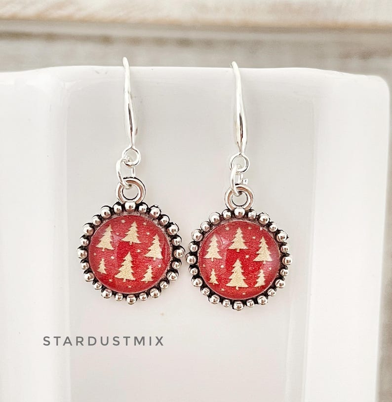 May include: A pair of silver-toned drop earrings featuring circular pendants with a red background and a repeating pattern of small white pine trees. The pendants are framed by a beaded silver border and hang from simple silver-toned ear wires.