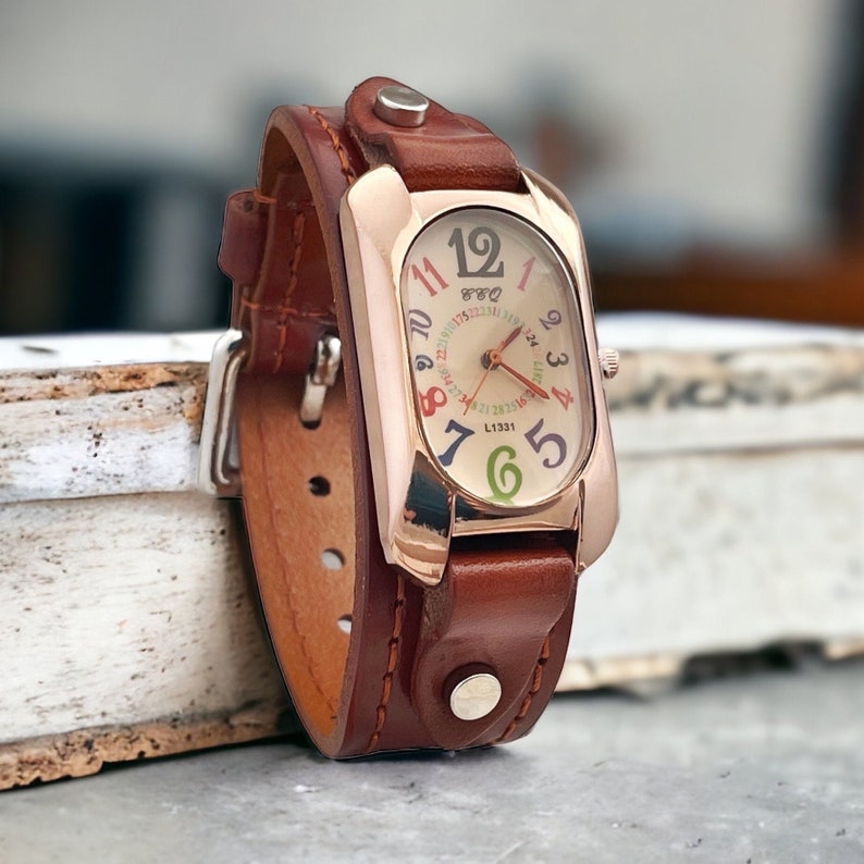 May include: A brown leather wristwatch with a rose gold case and a white face. The face has colourful numbers and a small second hand. The watch has a unique design with the leather strap wrapped around the case.