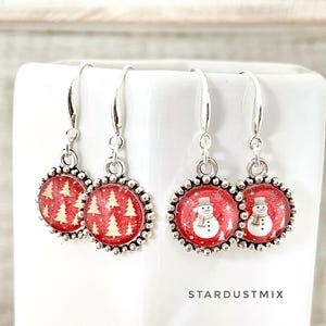 May include: Two pairs of silver-toned drop earrings featuring circular charms with a beaded border. One pair displays a red background with gold-colored pine trees, while the other pair shows a red background with white snowmen. Text reads STARDUSTMIX.
