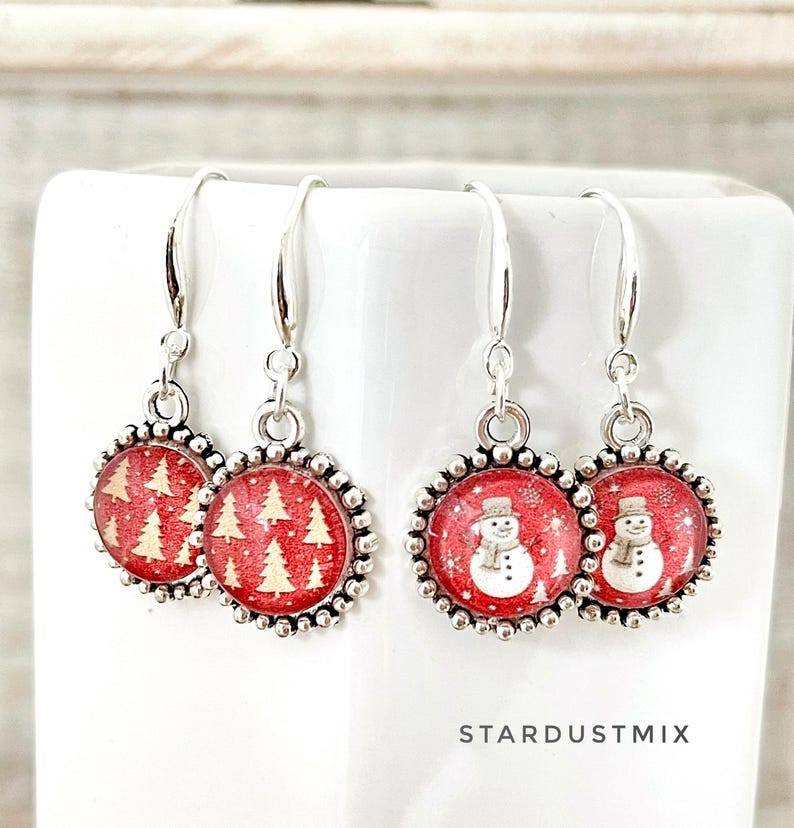May include: Two pairs of silver-toned drop earrings featuring circular charms with a beaded border. One pair displays a red background with gold-colored pine trees, while the other pair shows a red background with white snowmen. Text reads STARDUSTMIX.