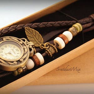 May include: A brown leather wristwatch with a gold-toned face and a leaf charm. The watch is adorned with brown leather straps and white, brown, and green beads.