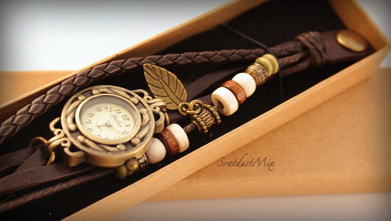 May include: A brown leather wristwatch with a gold-toned face and a leaf charm. The watch is adorned with brown leather straps and white, brown, and green beads.