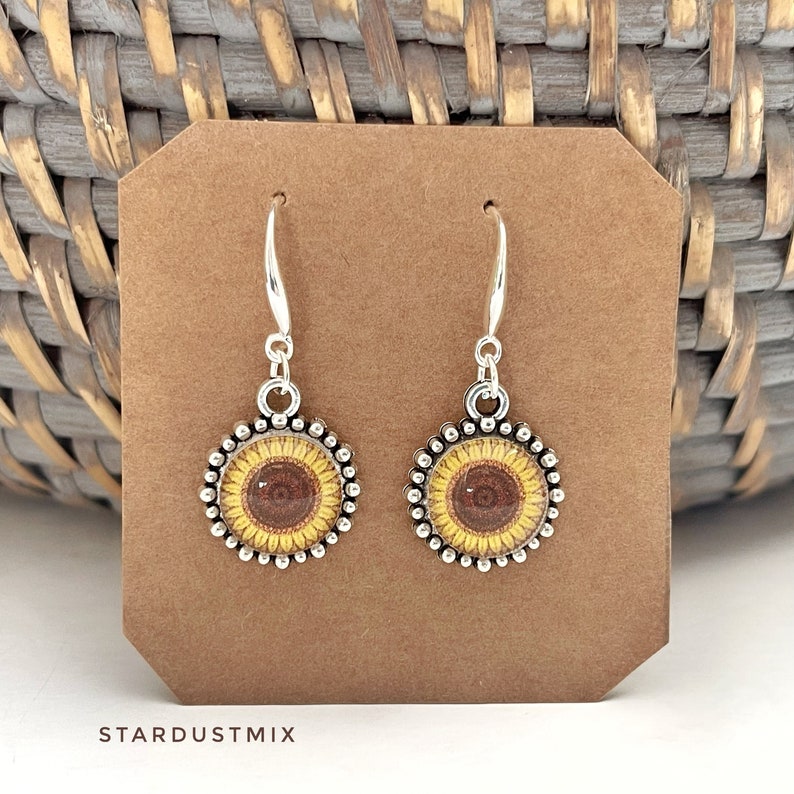 May include: A pair of silver-toned drop earrings featuring circular charms with a sunflower graphic. Each charm has a beaded edge and is suspended from a silver-toned hook. The text STARDUSTMIX is printed in black at the bottom of the display card.