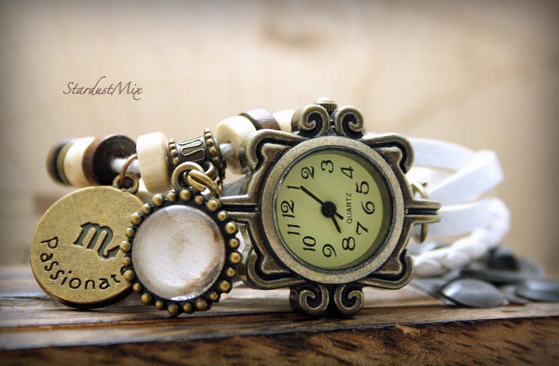 Puede incluir: Un reloj de cuero blanco con una esfera de color bronce y un dise&ntilde;o decorativo. El reloj tiene una esfera blanca con n&uacute;meros y manecillas negras. El reloj est&aacute; adornado con un brazalete de charms con una cuenta blanca, un charm de bronce con el s&iacute;mbolo astrol&oacute;gico de Virgo y la palabra "apasionado", y un peque&ntilde;o charm redondo blanco.