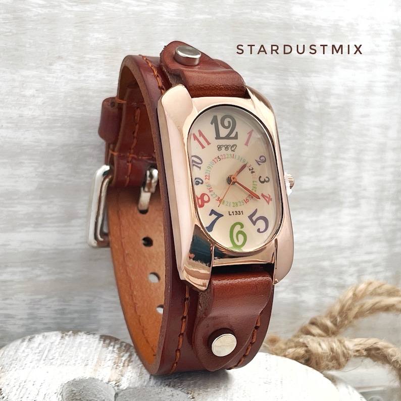 May include: A rose gold watch with a brown leather strap. The watch face has a white background with colourful numbers and a small calendar window. The watch face is oval shaped.