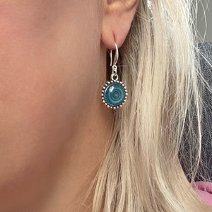 May include: A silver drop earring featuring a circular pendant with a blue and teal concentric circle pattern. The pendant is framed by a beaded silver border and hangs from a simple silver hook. The text STARDUSTMIX is visible at the bottom.