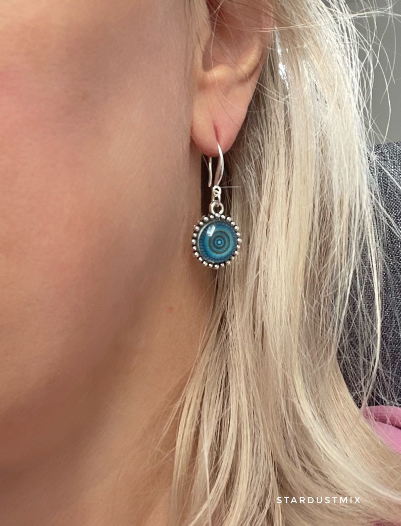 May include: A silver drop earring featuring a circular pendant with a blue and teal concentric circle pattern. The pendant is framed by a beaded silver border and hangs from a simple silver hook. The text STARDUSTMIX is visible at the bottom.