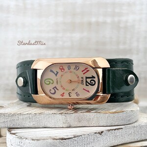 May include: A rose gold watch with a green leather strap. The watch face is oval shaped and has colourful numbers around the edge. The watch face also has the text "120 1910175 RO" printed on it.