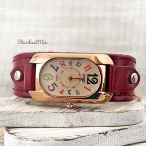 May include: A red leather cuff watch with a rose gold case and a white face with colourful numbers. The watch face has a date window and a small second hand.