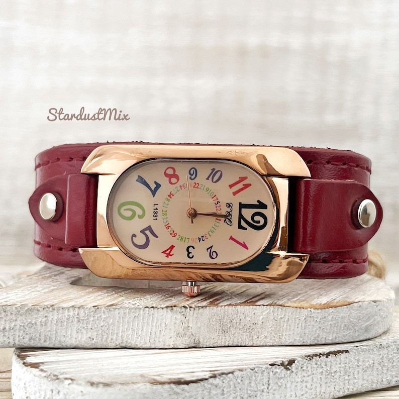 May include: A red leather cuff watch with a rose gold case and a white face with colourful numbers. The watch face has a date window and a small second hand.