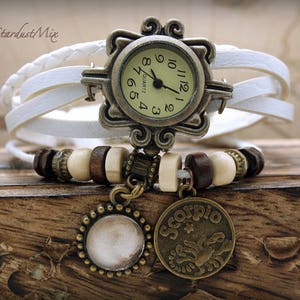 Puede incluir: Reloj de pulsera de cuero blanco con una esfera redonda dorada y agujas negras. El reloj tiene un marco decorativo adornado y un encanto con el s&iacute;mbolo del zod&iacute;aco de Escorpio.