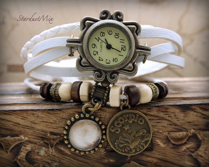 Puede incluir: Reloj de pulsera de cuero blanco con una esfera redonda dorada y agujas negras. El reloj tiene un marco decorativo adornado y un encanto con el s&iacute;mbolo del zod&iacute;aco de Escorpio.