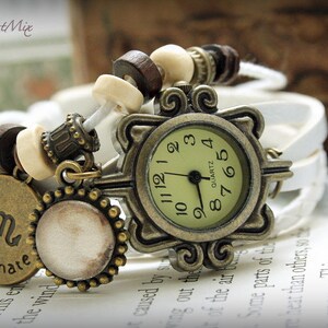 Puede incluir: Un reloj de pulsera de cuero blanco con una esfera dorada y cuentas de madera marr&oacute;n. La esfera del reloj tiene marcas de n&uacute;meros romanos y las palabras "QUARTZ" y "8765". El reloj est&aacute; decorado con un amuleto que dice "apasionado" con el s&iacute;mbolo astrol&oacute;gico de Escorpio.