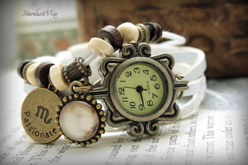 Puede incluir: Un reloj de pulsera de cuero blanco con una esfera dorada y cuentas de madera marr&oacute;n. La esfera del reloj tiene marcas de n&uacute;meros romanos y las palabras "QUARTZ" y "8765". El reloj est&aacute; decorado con un amuleto que dice "apasionado" con el s&iacute;mbolo astrol&oacute;gico de Escorpio.