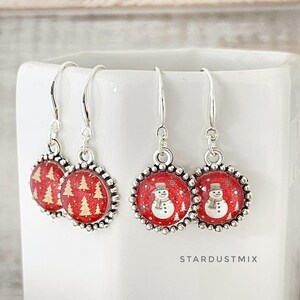 May include: Two pairs of silver-toned drop earrings featuring circular charms with a red glitter background. One pair displays gold-colored pine trees, while the other pair features white snowman illustrations. The text STARDUSTMIX is visible.