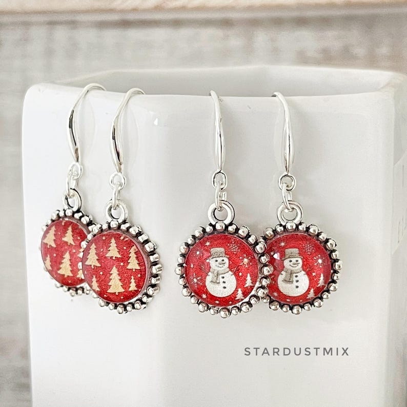 May include: Two pairs of silver-toned drop earrings featuring circular charms with a red glitter background. One pair displays gold-colored pine trees, while the other pair features white snowman illustrations. The text STARDUSTMIX is visible.