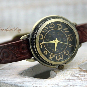 Könnte beinhalten: Vintage Armbanduhr mit dunklem Zifferblatt und goldfarbenen Zeigern und Ziffern. Die Uhr hat ein braunes Lederarmband mit geprägtem floralem Design. Das Zifferblatt zeigt den Text "Stardust Mix".