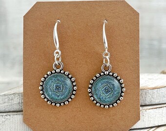May include: A pair of silver dangle earrings with blue and green mandala designs. The earrings are on a brown card with the text "Stardustmix" printed on it.